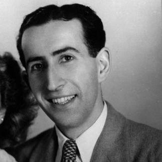 Black-and-white studio photograph of a man sitting and smiling at the camera. The man wears a suit and has dark brown hair.”