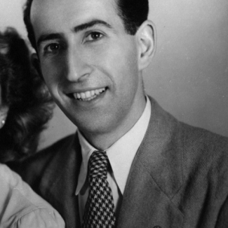 Photo en noir et blanc prise en studio d'un homme assis, souriant à la caméra. Il a des cheveux bruns foncés.”