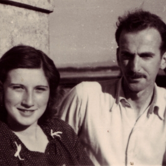 Photo en noir et blanc d'un homme et d'une femme, assis ensemble à l'extérieur. La femme a de cheveux bruns jusqu'aux épaules et l'homme porte une moustache ainsi qu'une chemise à col de couleur pâle.