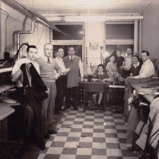 Black-and-white photograph of a group of about ten people standing or sitting together in a room with two windows at the back. The group looks at the camera. Piles of fabric and clothing are on the right, and the floor is checkered.