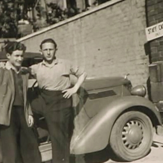 Black-and-white photograph of two young men standing together in front of a vintage car outdoors. One man has his arm around the other. A large brick wall is in the background.