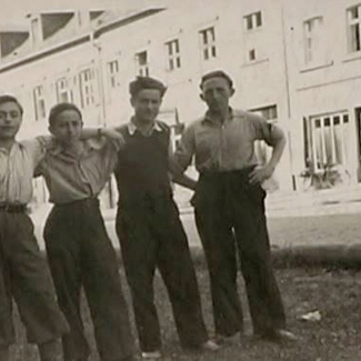 Black-and-white photograph of a group of four boys standing together arm-in-arm outdoors, in a shaded area, on a road. Across the street is a large building.