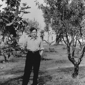 Black-and-white photograph of a young man standing on a lawn, with a tree on either side of him. He has his hands in his pockets, and wears dark trousers and a light collared shirt.