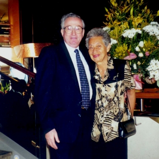 Photo en couleur d'un couple de personnes âgées se tenant ensemble bras-dessus bras-dessous et souriant. L'homme porte un complet et la femme porte une blouse à motifs. Il sont debout devant un grand arrangement floral.