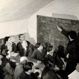 Black-and-white photograph of a group of about a dozen men looking at a chalk board. One of the men stands with a piece of chalk and points at an equation written on the board. The other men are sitting at a table, some taking notes with pen and paper.