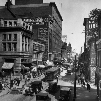 Black-and-white photograph of a busy street scene with vintage cars and streetcars driving up and down a road. Pedestrians walk in either direction on the sidewalks, and there are billboard advertisements posted on buildings’ exterior walls.