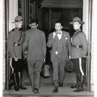 Photo en noir et blanc de quatre hommes dans une grande porte. Un homme, le deuxième à partir de la droite, sort de la pièce avec un attaché-case et a une pipe dans sa bouche. L'homme à sa gauche entre dans la pièce. Les deux hommes se tenant de part et d'autre portent un uniforme de la Gendarmerie royale du Canada et regardent en direction de la caméra.