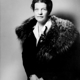 Photo en noir et blanc d’un portrait de femme à partir de la taille. Elle porte un manteau noir avec un col de fourrure. Ses cheveux sont courts et frisés et elle regarde directement la caméra.