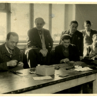 Black-and-white photograph of a group of six men, three standing and three sitting at a table. The men standing are observing the sitting men as they sew fabric with a needle and thread at a table.