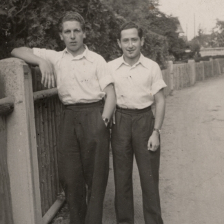 Black-and-white photograph of two young men standing together on a road, one of them leaning against a fence. They wear white collar, short sleeved shirts, and dark trousers.