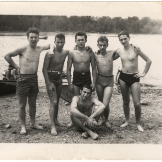 Photo en noir et blanc d'un groupe de six adolescents se tenant ensemble bras-dessus bras-dessous sur une plage avec de l'eau derrière eux. Les jeunes hommes portent des maillots de bain et l'un d'eux est assis au sol.