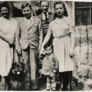 Photo en noir et blanc d'un homme et d'une femme debout à l'extérieur d'une maison avec deux adolescents, un jeune enfant et un chien.