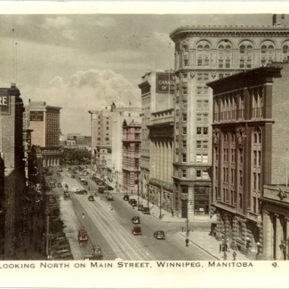 Photo en noir et blanc de bâtiments légèrement colorés en jaune et rose, représentant la vue d'une rue d'une ville où il y a des édifices en brique et des vitrines de commerces. Il y a des affiches sur trois bâtiments pour The McIntyre Block, Marathon Blue Gas et Bank of Montreal.