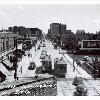 Photo en noir et blanc d'une scène d'une rue, prise en hauteur. Une large rue où circulent des piétons sur les trottoirs, il y a des vitrines de commerce de chaque côté, et trois tramways qui circulent au centre de la rue.