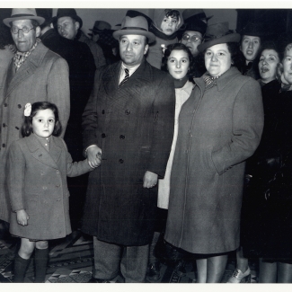 Black-and-white photograph of a group of about a dozen people standing together and looking at the camera. They wear coats and hats. The man in the front row holds the hand of a young girl with a bow in her hair.