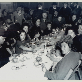 Photo en noir et blanc d'un grand groupe de personnes assis ensemble à longue table, regardant vers la caméra. La table est remplie de plats et de nourriture. Il y a plusieurs personnes dans la pièce derrière la table.