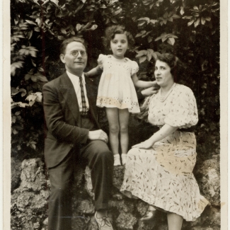 Photo en noir et blanc d'un homme et d'une femme assis à l'extérieur sur un mur de pierre avec une jeune fille debout entre eux, s'appuyant sur leurs épaules. La femme et sa fille portent des robes et l'homme un complet.