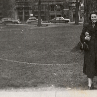 Photo en noir et blanc d'une femme souriant et s'appuyant sur un lampadaire devant un grand champ avec des bâtiments et des voitures en arrière-plan. Elle tient un sac-à-main et un livre dans sa main.