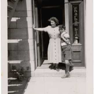 Photo en noir et blanc d'une femme et d'un garçon se tenant debout à la porte d'entrée d'un appartement du rez-de-chaussée. La femme tient son fils d'un bras et s'appuie sur le cadre de la porte de l'autre.