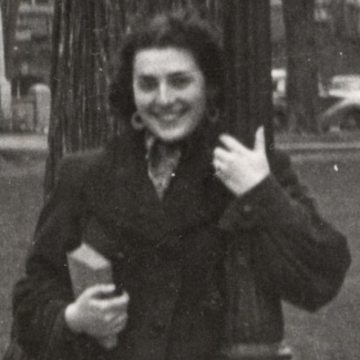 Photo en noir et blanc d'une femme souriant et s'appuyant sur un lampadaire devant un grand champ avec des bâtiments et des voitures en arrière-plan. Elle tient un sac-à-main et un livre dans sa main.