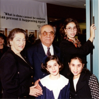 Colour photograph of an elderly man and woman, standing together indoors with a woman and two young girls. The family is formally dressed, with the girls wearing blouses and the man in a suit.