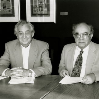 Black-and-white photograph of two elderly men sitting at a table with papers in front of them. The men wear suit jackets, one them wearing a tie, and there are three framed artworks hung on a wall in the background.