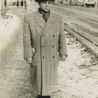 Black-and-white photograph of a man standing outdoors on a snowy sidewalk. The man wears a hat and coat, and there are buildings in the background.