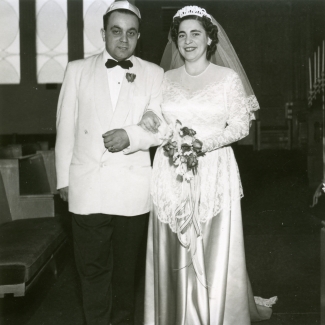 Photo en noir et blanc d'un homme et d'une femme se tenant à l'intérieur bras-dessus bras-dessous. Le couple semble célébrer leur mariage. L'homme porte un complet avec un veston de couleur claire et un nœud papillon, et la femme une longue robe blanche dont la jupe est en soie, un voile et elle tient un bouquet de fleurs.