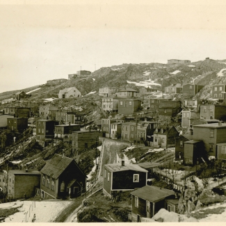 Black-and-white photograph of small residential buildings in a hillside town or village. There is snow on the ground and it appears to be winter.