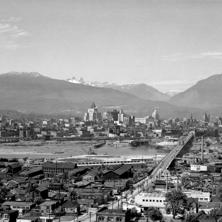 Photo en noir et blanc, vue aérienne d'une ville. Plusieurs bâtiments sont répartis de part et d'autre d'une rivière. Un pont reliant les deux rives se trouve dans la partie droite de la photo. Plusieurs sommets montagneux sont visibles à l'arrière-plan.