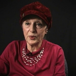 Screenshot of Holocaust survivor Sarah Engelhard video testimony. She is sitting in front of a black background, and looking to the left of the camera. The camera shows her face and shoulders.