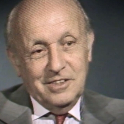 Screenshot of Holocaust survivor George Lysy video testimony. He is sitting in front of a grey background, and looking to the left of the camera. The camera shows his face and shoulders.