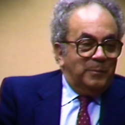 Screenshot of Holocaust survivor David Shafran video testimony. He is sitting in front of a beige background, and looking to the right of the camera. The camera shows his face and shoulders.