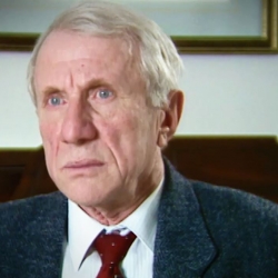 Screenshot of Holocaust survivor Elliott Zuckier video testimony, sitting in his living room, and looking to the left of the camera. The camera shows his face and shoulders.